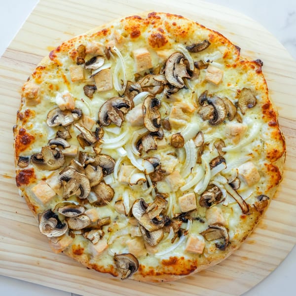 A RigaTony’s white pizza with chicken, mushrooms, and onions on a wooden pizza peel.