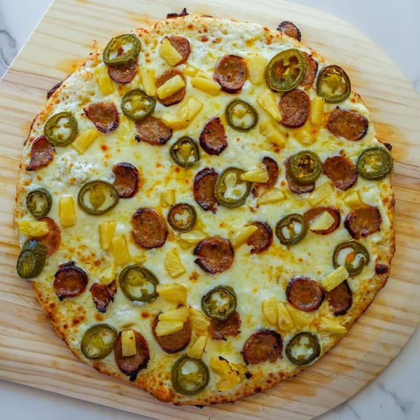 RigaTony's pizza with white sauce, sausage, pineapple, and jalapeños on a wooden peel.