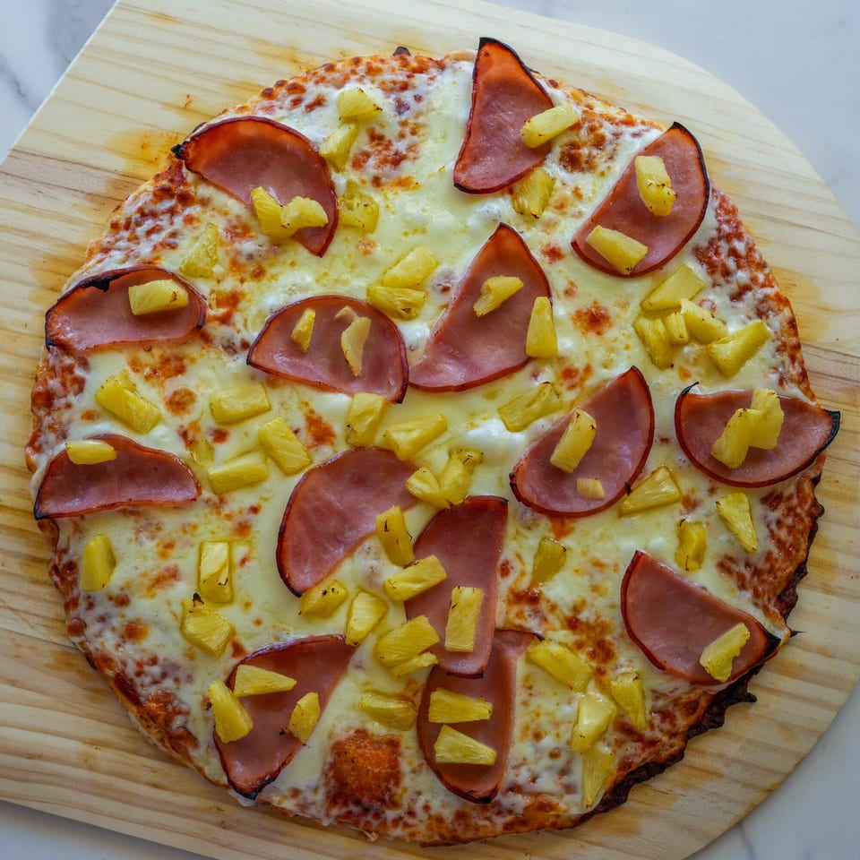 A whole RigaTony's Hawaiian pizza with ham and pineapple on a wooden paddle.