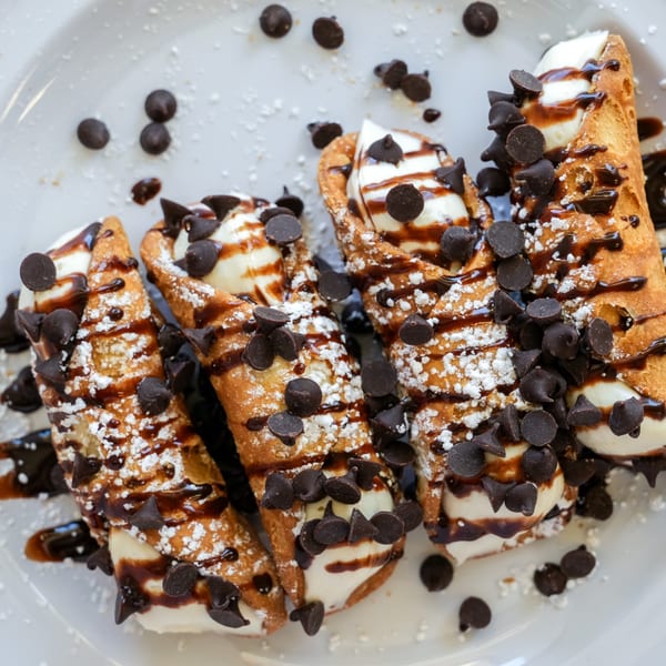 Four cannolis filled with cream, drizzled with chocolate syrup, and topped with chocolate chips and powdered sugar.
