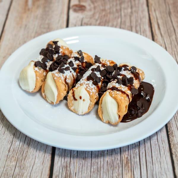 A plate of RigaTony's cannolis, drizzled with chocolate sauce, chocolate chips, and powdered sugar.