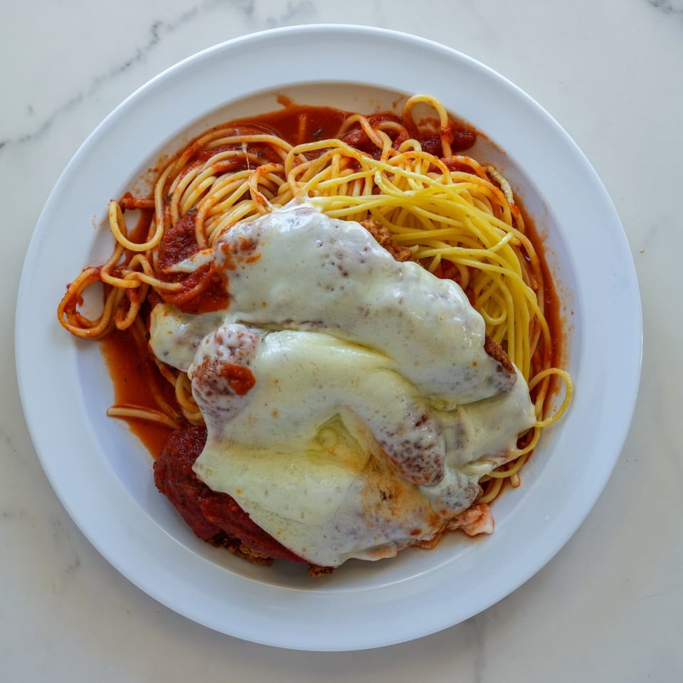 A white plate of RigaTony's spaghetti with meatballs and melted cheese in red sauce.