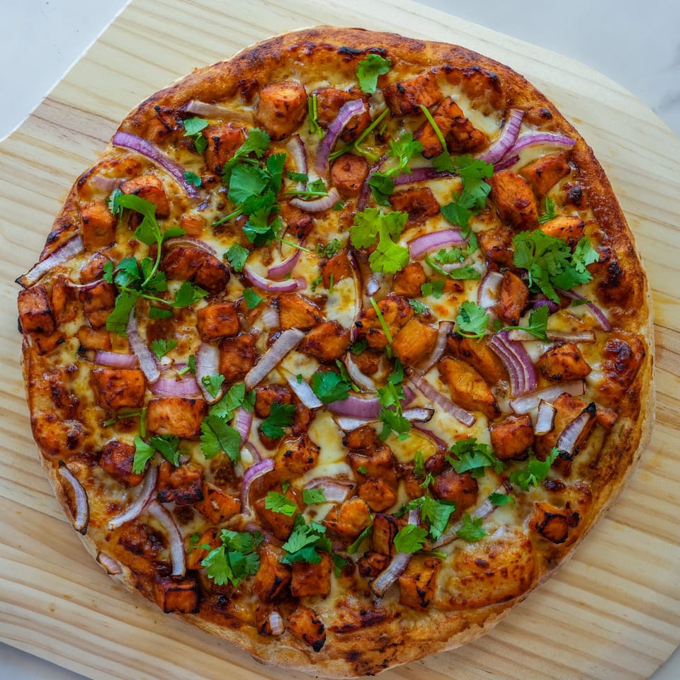 A RigaTony's pizza topped with chicken, red onions, and cilantro on a wooden board.