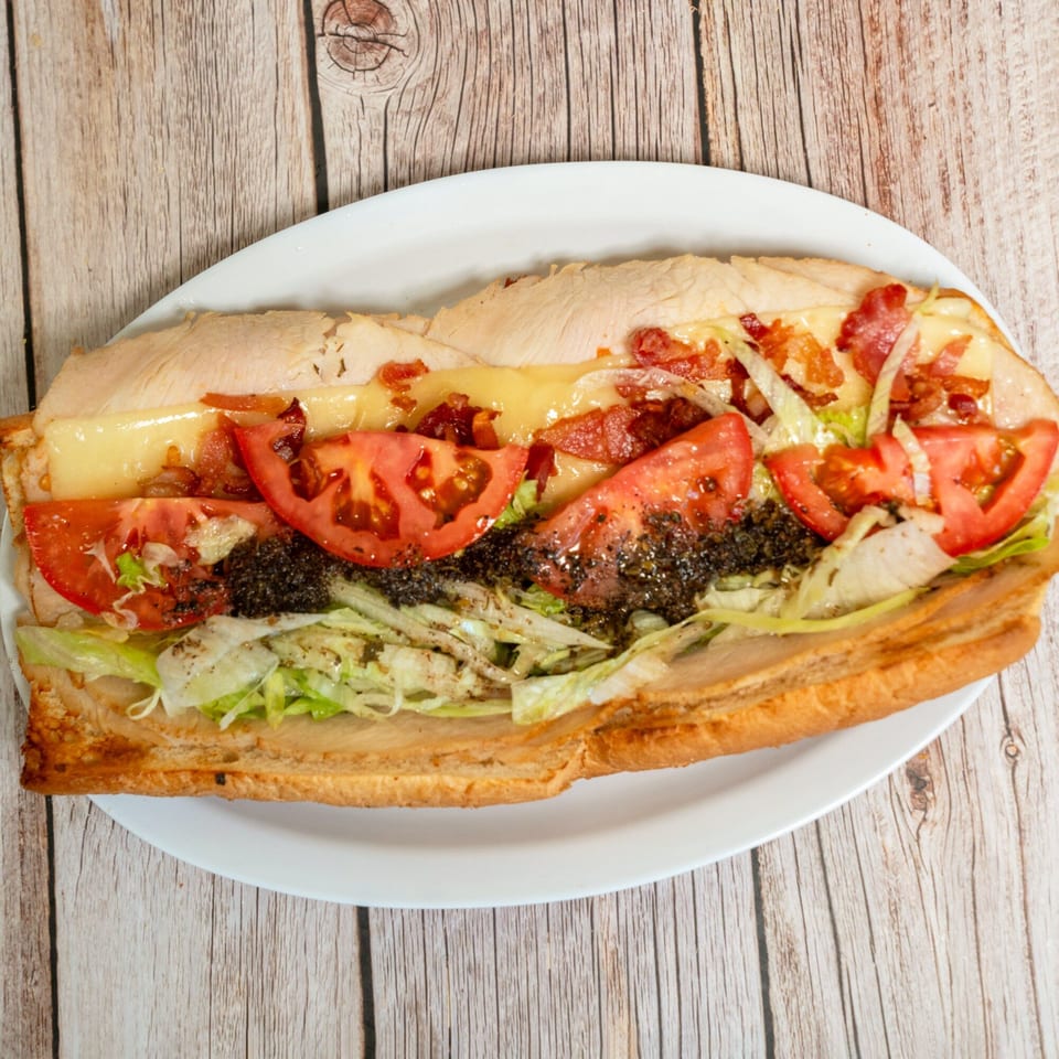 A RigaTony’s sub sandwich with turkey, bacon, cheese, tomatoes, lettuce, and pesto on a white plate.